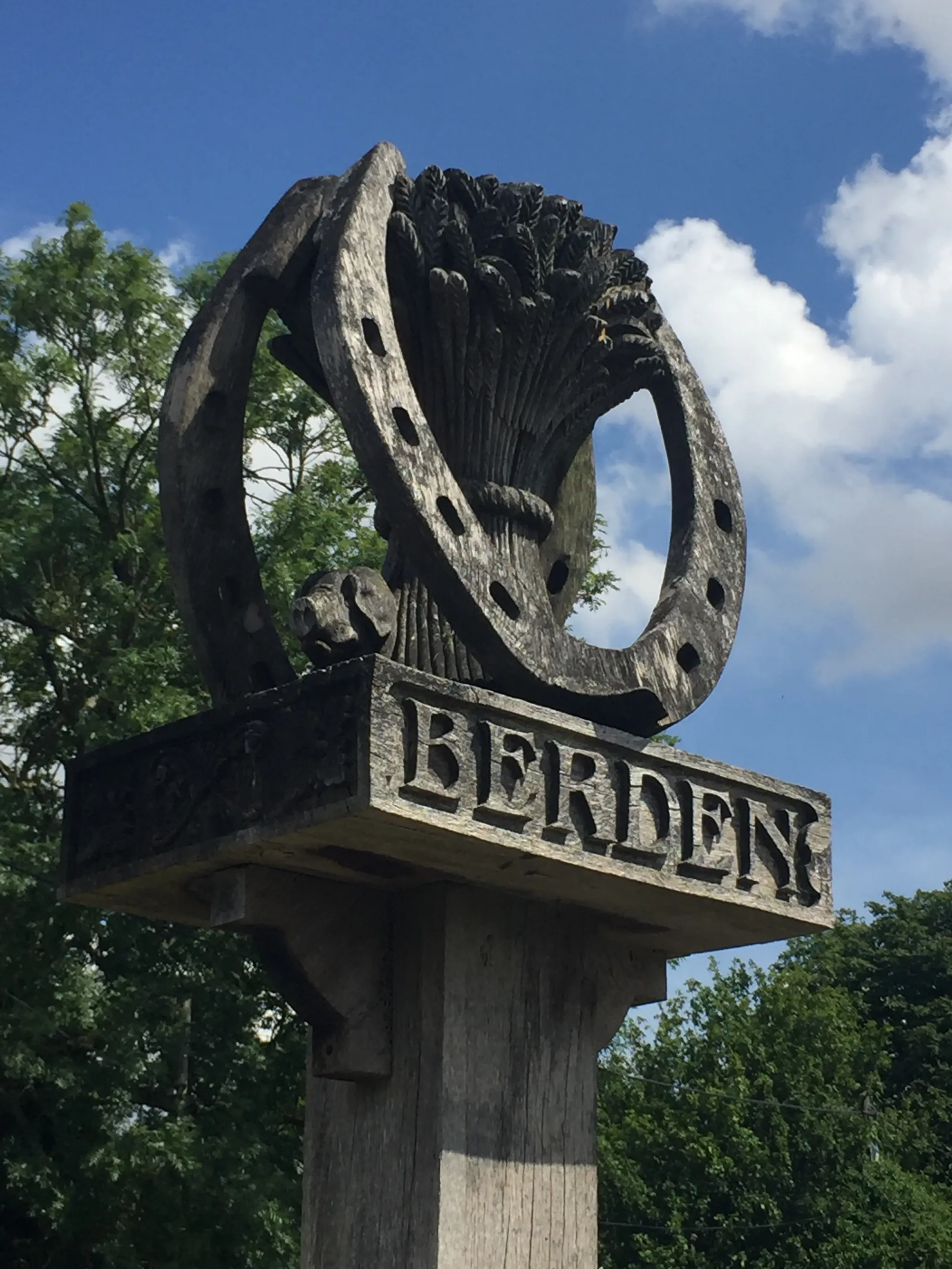 Wooden signpost for the village of Berden, with carved wheat sheaf and horseshoe symbol above the inscription, against a partly cloudy blue sky, with trees in the background.