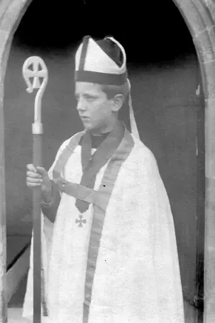 A young boy dressed in bishop's attire, wearing a mitre hat and holding a crosier, standing in a stone archway.