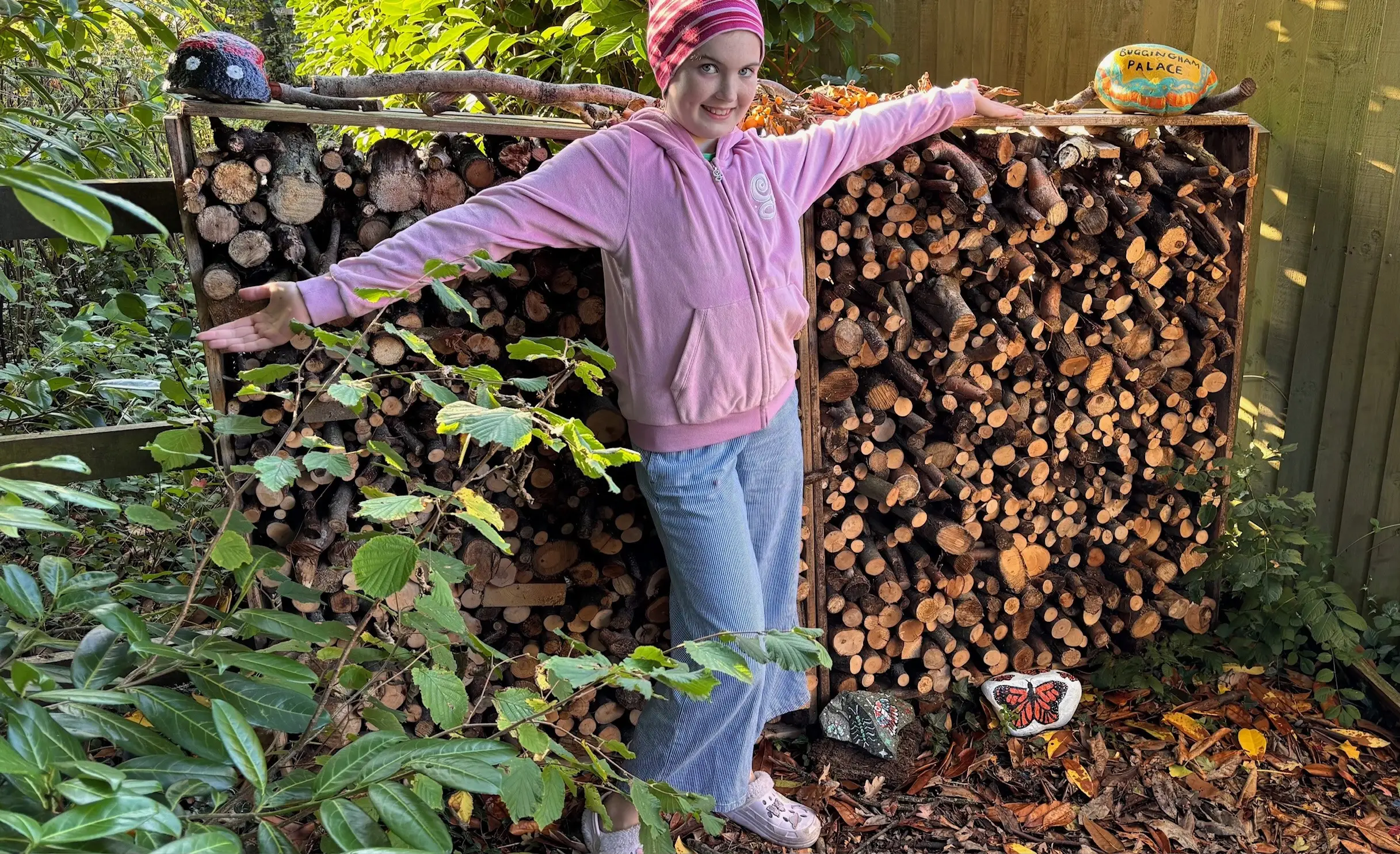 A young woman with a pink hoodie and striped beanie poses with arms outstretched in front of a stacked woodpile, smiling at the camera, with a painted rock saying "Buggingham Palace" above, a painted butterfly rock at her feet, and a whimsical decorative wooden hedgehog on top.