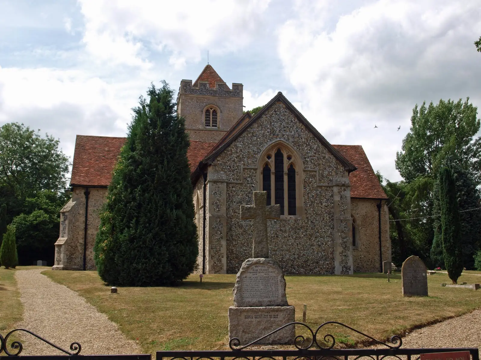 A view with the Berden War Memorial cross backed by St Nicholas Church viewed from a gravel path lined with trees and tombstones under a cloudy sky. 